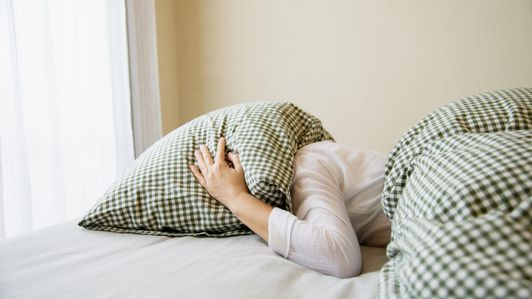 Young Woman Covering Pillow Lying On Bed Against Wall At Home